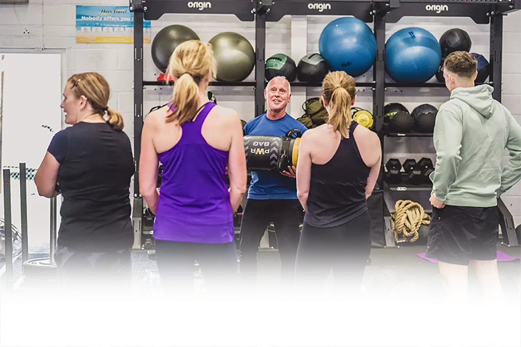 Trainer demonstrates a weighted sandbag lift to a small group in a gym with rack of medicine balls behind them