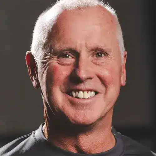 Smiling middle-aged man with short white hair looking at the camera in a neutral backdrop.