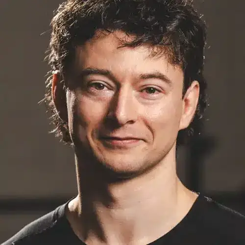 Young man with short dark hair and a subtle smile, wearing a black shirt against a plain background.