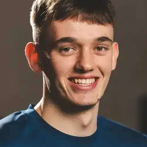 Head-and-shoulders portrait of a young man with short dark hair, smiling at the camera, wearing a blue shirt.