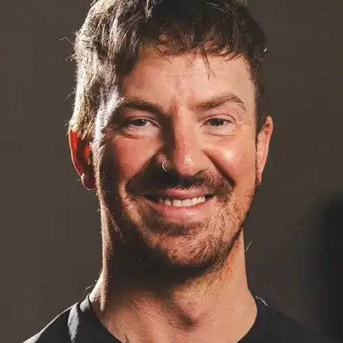 Portrait of a smiling man with short dark hair and light stubble, wearing a dark shirt and looking at the camera.